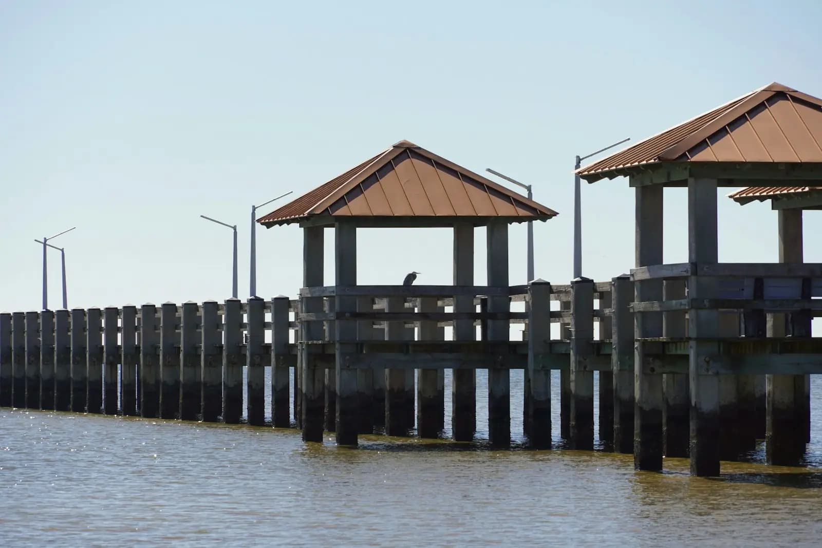 A long low fishing pier with covered shelters standing over flat brown water under a pale sky
